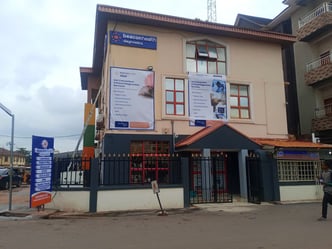 Exterior of MDaaS Global's new BeaconHealth diagnostics centre in Enugu, Nigeria — a two-storey building with dark and cream façade, red-framed windows, and BeaconHealth signage and service boards along the front
