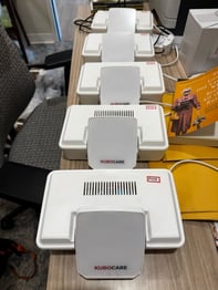 Several white Kubo Care fall-detection sensor units lined up on a wooden desk, ready for installation in senior living communities.