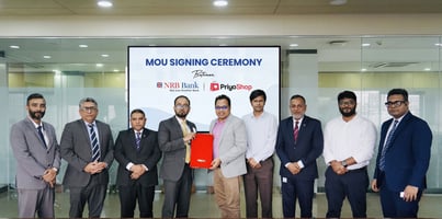 Leaders from PriyoShop and NRB Bank PLC stand in a row holding the signed agreement in front of a screen reading “MOU Signing Ceremony between NRB Bank and PriyoShop
