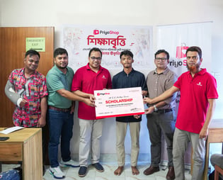 Priyoshop team standing together at a PriyoShop event, presenting an oversized scholarship certificate to students. A backdrop behind them reads 'শিক্ষাবৃত্তি' (scholarship) alongside the PriyoShop logo