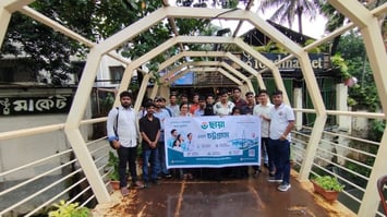 A Chhaya team of around a dozen people standing under an arched wooden walkway in Chittagong, holding a branded banner in Bengali announcing the launch of Chhaya's services in the city