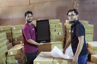 Two men in a warehouse holding up a ChutneyAds digital display screen, surrounded by stacks of packaged screens ready for rollout to PriyoShop's MSME retail network